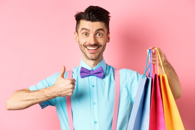Счастливый мужчина-покупатель показывает палец вверх и хозяйственные сумки, рекомендуя магазин, стоя на розовом фоне.