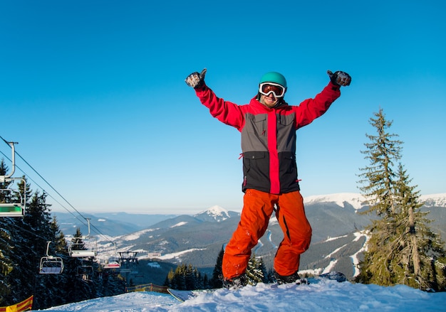 幸せな男性スノーボーダーが山の冬のスキーリゾートの雪の斜面に立って、親指を現して。スキー場のリフトと青い空