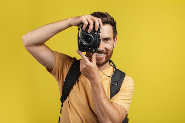 Happy male tourist with backpack taking picture on professional DSLR camera over yellow studio background