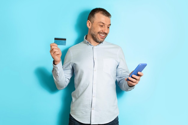 Счастливый мужчина с помощью смартфона и кредитной карты делает покупки на синем фоне