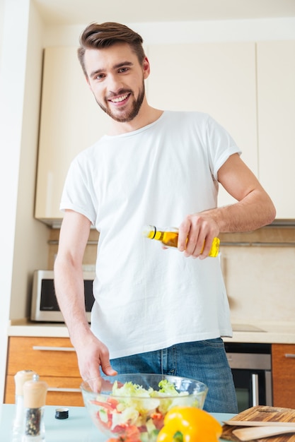 Uomo felice che aggiunge olio d'oliva in insalata in cucina e guarda davanti