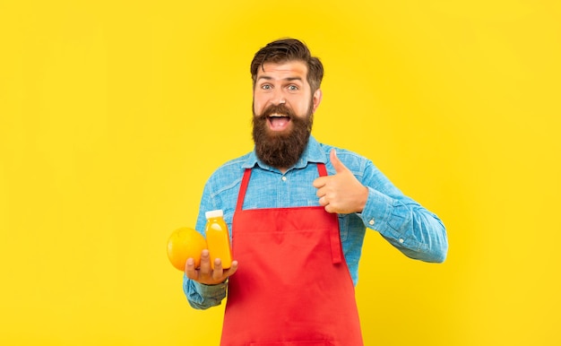 Счастливый человек в фартуке, держащий большой палец, держащий апельсин и бутылку сока, владелец магазина сока