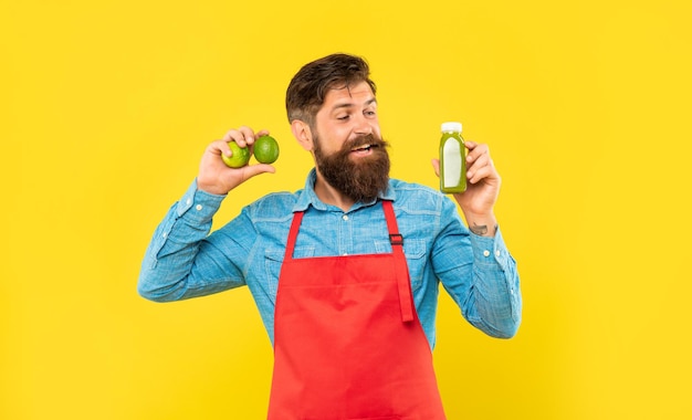 Happy man in apron looking at juice bottle holding limes yellow background juice barman