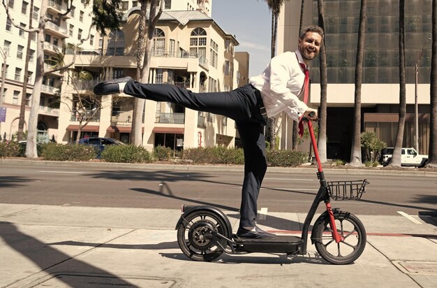 Happy man businessman in suit riding electro scooter outdoor on cityscape urban background rental transportation