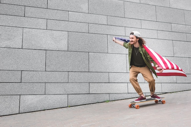 Uomo felice in abbigliamento casual con bandiera americana in sella a skateboard