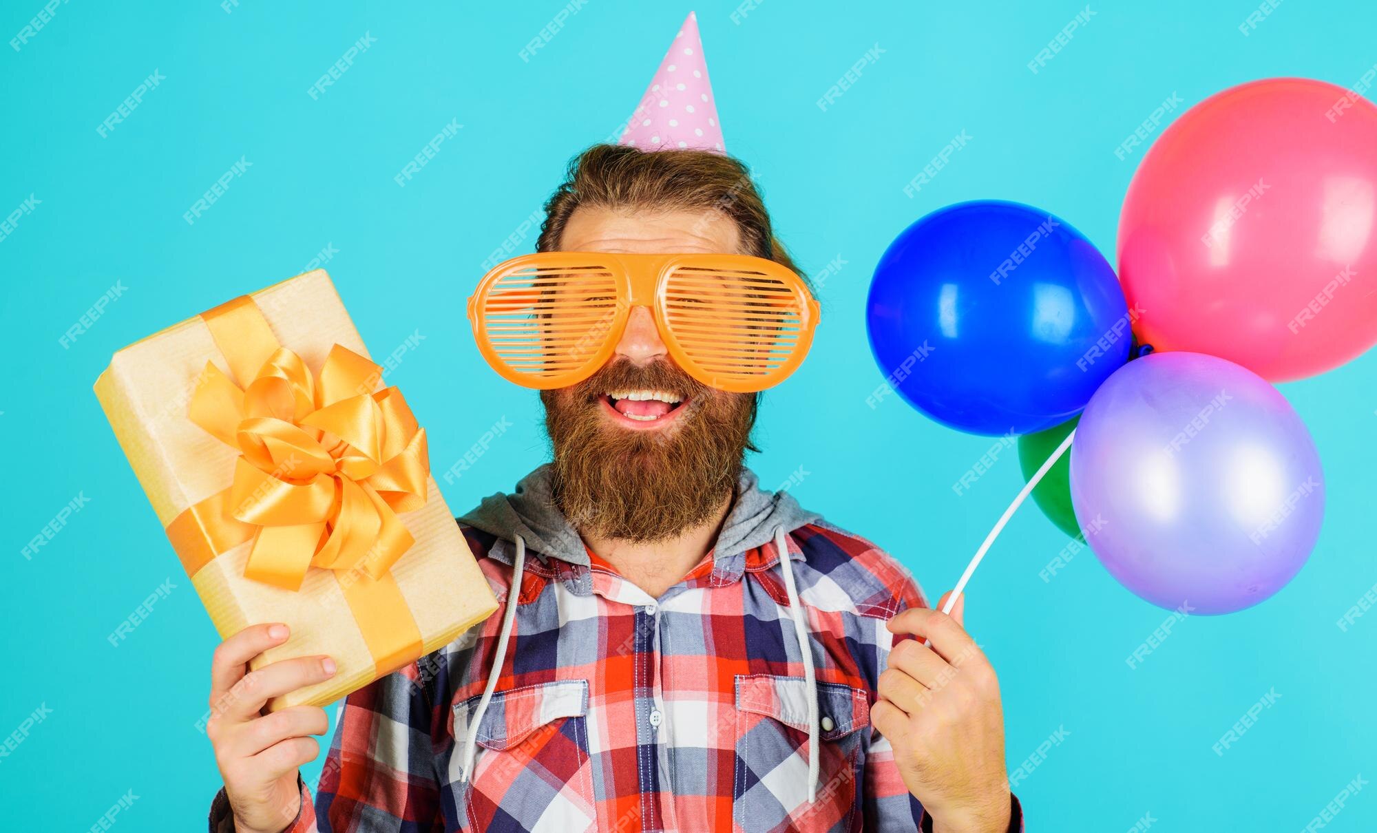 風船とギフトボックスのパーティータイムでホリデーキャップで誕生日のひげを生やした男を祝う幸せな男 プレミアム写真