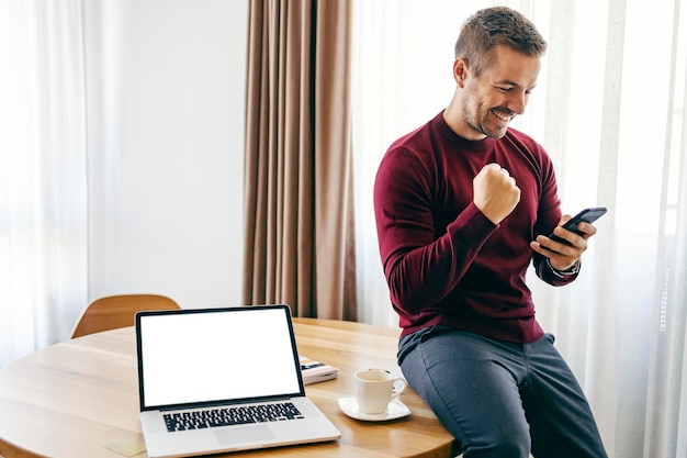 A happy man celebrating success while sitting on the table at home and smiling at the phone