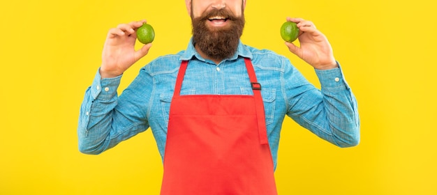 Happy man crop view in red apron holding fresh limes citrus fruits yellow background fruiterer