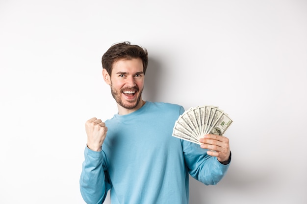 Happy man dancing with money, showing dollars and saying yes with satisfied smile, making fist pump gesture, standing over white background