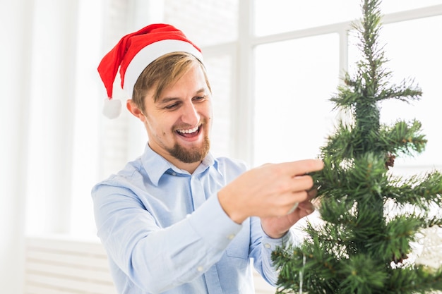 Uomo felice che decora l'albero di natale a casa con il cappello di babbo natale. uomo che decora l'albero con le palline durante le vacanze invernali.