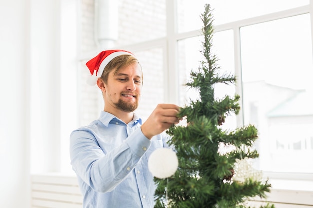 サンタクロースの帽子で自宅でクリスマスツリーを飾る幸せな男。冬の休暇中につまらないもので木を飾る男。