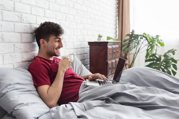 Happy man drinking coffee and using laptop in bed in the morning