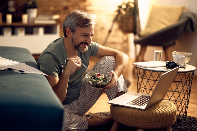 Uomo felice che mangia insalata mentre naviga in rete a tarda notte a casa