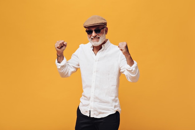 Happy man in fashionable outfit poses on orange background Handsome adult guy with gray beard in beige cap is happy and smiling