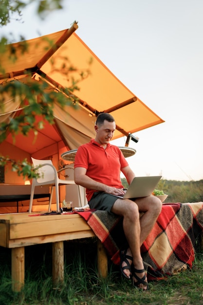 Happy man freelancer programmer using a laptop on a cozy glamping tent in a summer day Luxury camping tent for outdoor holiday and vacation Lifestyle concept