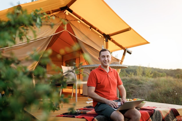 여름날 아늑한 글램핑 텐트에서 노트북을 사용하는 행복한 남자 프리랜서 야외 휴가 및 휴가 라이프스타일 컨셉을 위한 럭셔리 캠핑 텐트