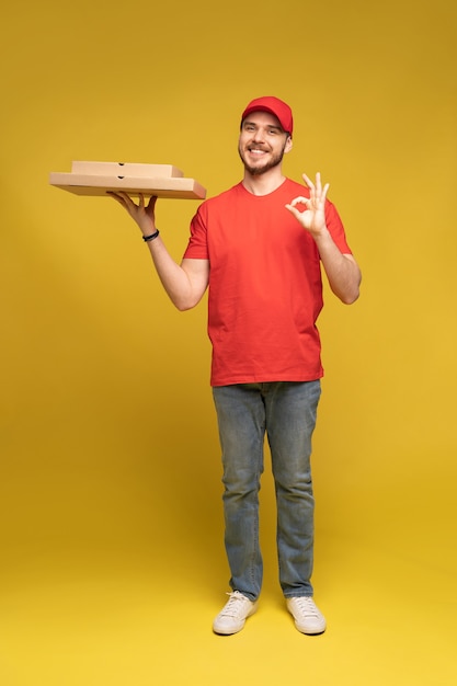 赤いTシャツとキャップで配達サービスから幸せな男が食べ物を注文し、黄色の壁に隔離されたピザボックスを保持しています