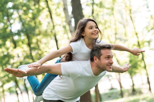 Photo happy man giving piggyback to his beautiful daughter with arms outstretched in park