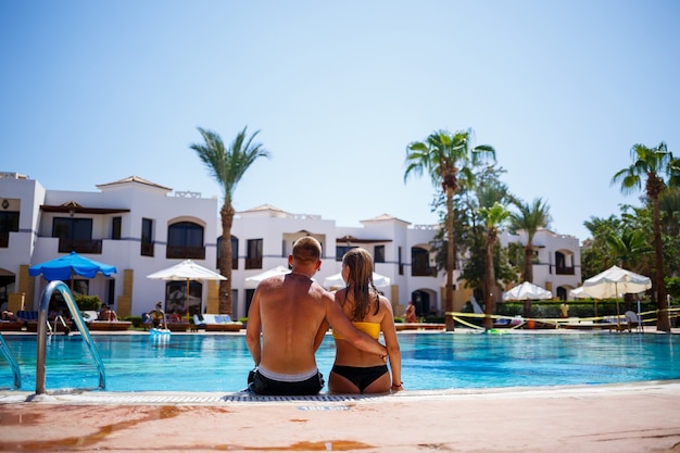 Happy man guy joy on vacation, girl sitting next to in a yellow swimsuit. Large pool, palm trees, blue water in the pool