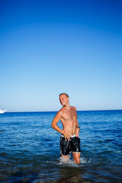 Happy man guy joy on vacation, stands at sea and splashes in the water. Men's shorts, sun, beach