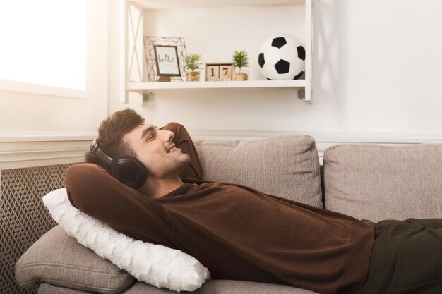 Happy man in headphones lying on couch at home. Dreaming and relaxing, listening to favorite music, copy space