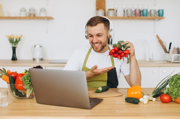 Happy man in headphones sitting at kitchen table and preparing salad holding radish and showing it to laptop