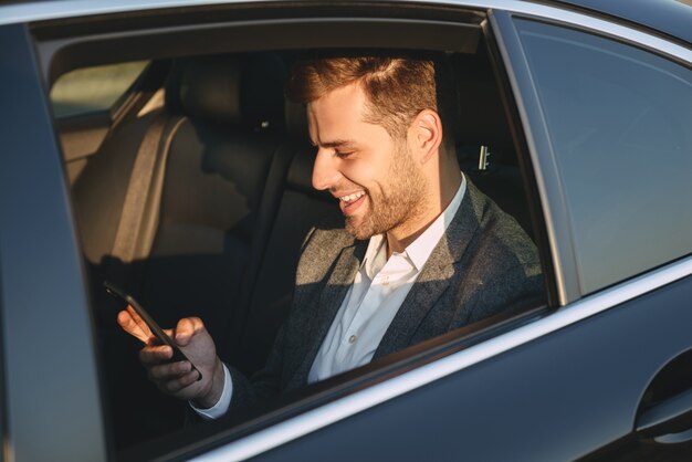 ビジネスクラスの車に戻って座っている間、携帯電話を保持し、使用して古典的なスーツで幸せな男