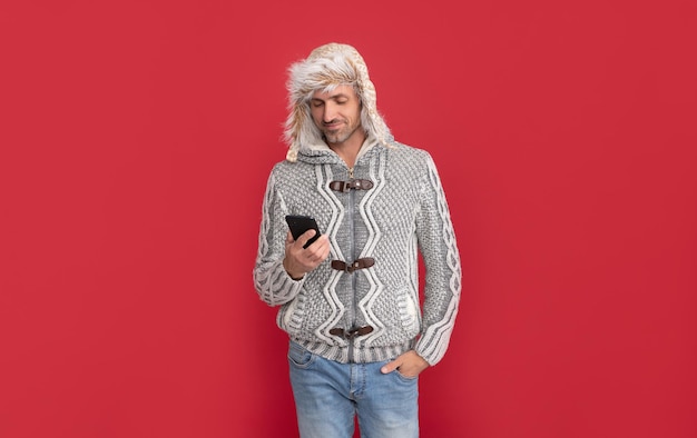 Фото Счастливый мужчина в свитере и ушанке проверяет электронную почту по телефону на красном фоне онлайн