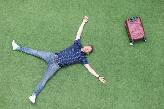 Photo happy man lies on green lawn with suitcase top view