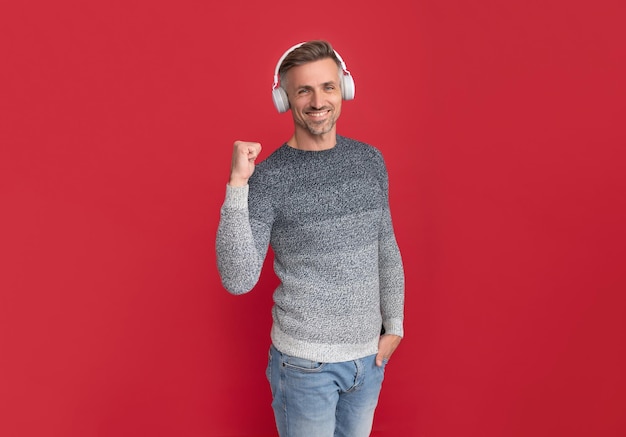 Happy man listen to music in headphones making winning gesture red background listening