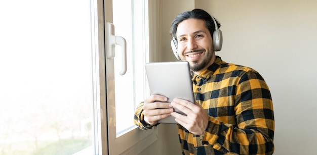 Happy man looking at the camera while holding a tablet with both hands and wearing headphones