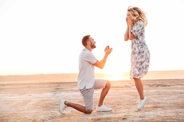 Uomo felice che fa proposta alla sua donna eccitata con anello in confezione regalo mentre cammina sulla spiaggia soleggiata