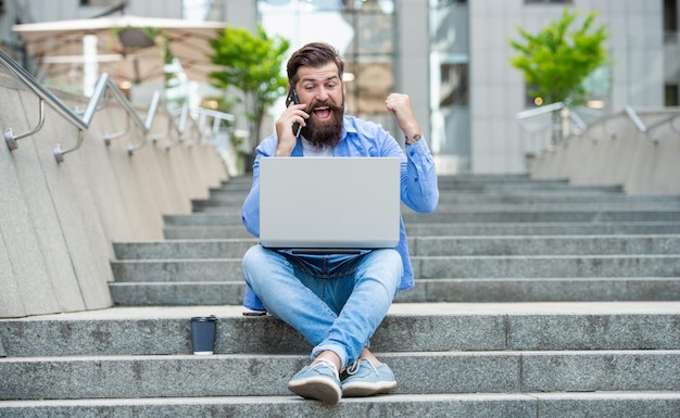 Happy man making winning gesture talking on mobile phone Guy celebrating betting win sitting on stairs Betting online Mobile betting Betting to win