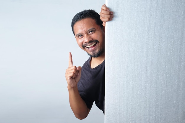 Photo happy man peeking behind the wall while pointing finger up