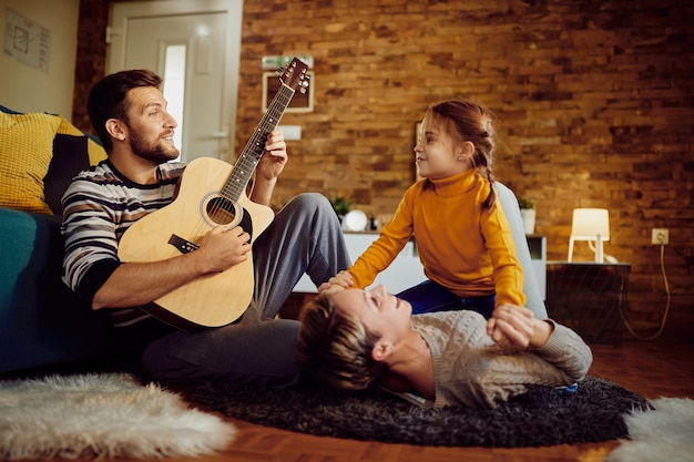 Happy man playing music on acoustic guitar while enjoying with his wife and daughter at home