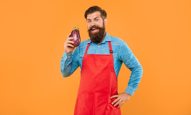 Happy man in red apron holding eggplant yellow background grocer