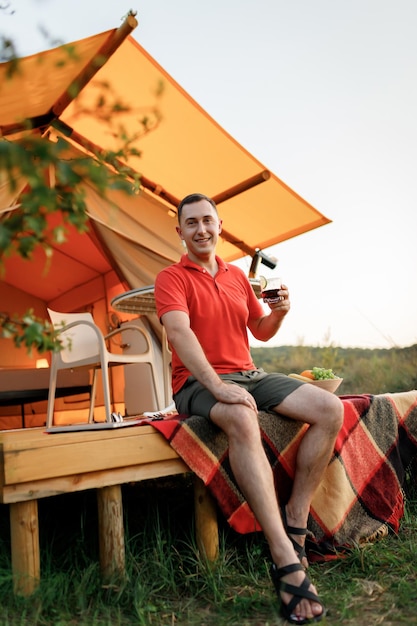 Happy man relaxing in glamping on summer evening drinking wiskiey with cola Luxury camping tent for outdoor recreation and recreation Lifestyle concept