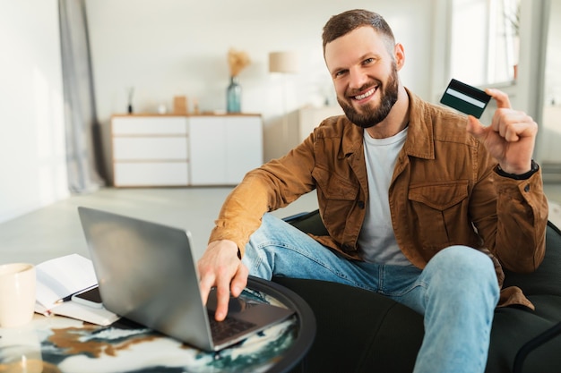 Happy man shopping online via laptop showing credit card indoor