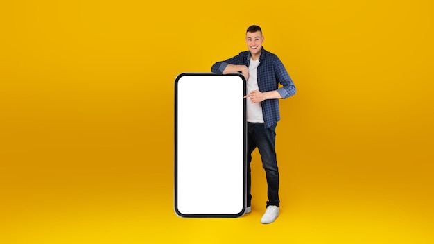 Happy man showing huge phone screen pointing fingers yellow background