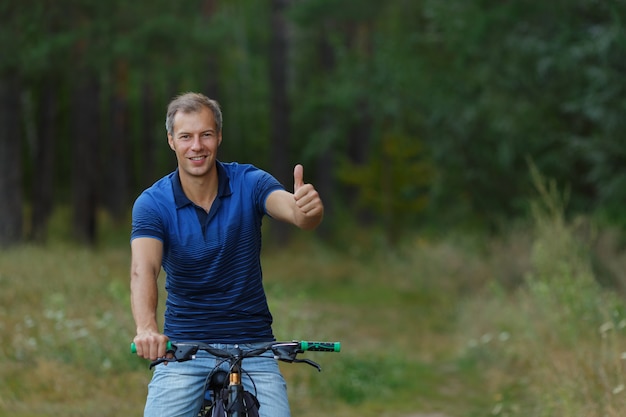 Happy man shows thumb up, riding on bicycle in pine forest, sport leisure. Cyclist walk in park, active healthy lifestyle