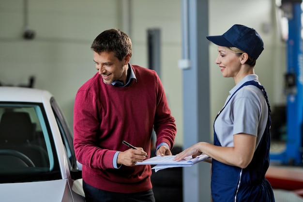 Happy man signing paperwork while talking to female mechanic in auto repair shop