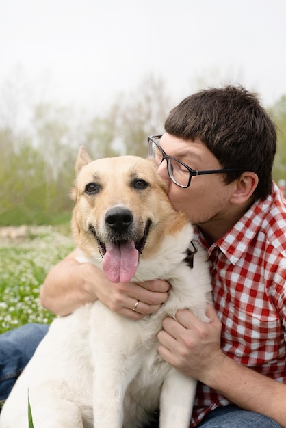 春の花の緑の草の上に混合品種の羊飼いの犬と一緒に座っている幸せな男