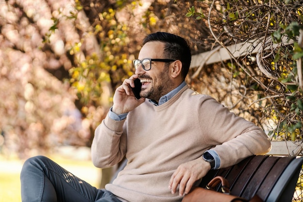 A happy man in smart casual is talking on the phone while sitting on a bench in park