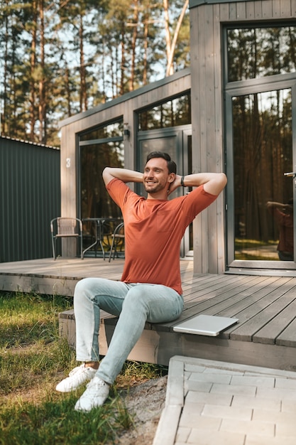 幸せな男。晴れた日の午後に家のポーチでラップトップの近くに座っているTシャツとジーンズで幸せなストレッチ男を笑顔