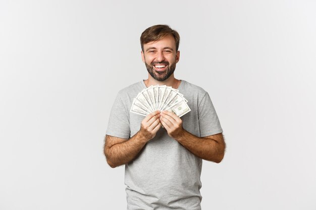 Счастливый человек улыбается довольным, показывая деньги, стоя на белом фоне в серой футболке