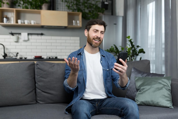 Happy man smiling and using phone sitting on sofa at home