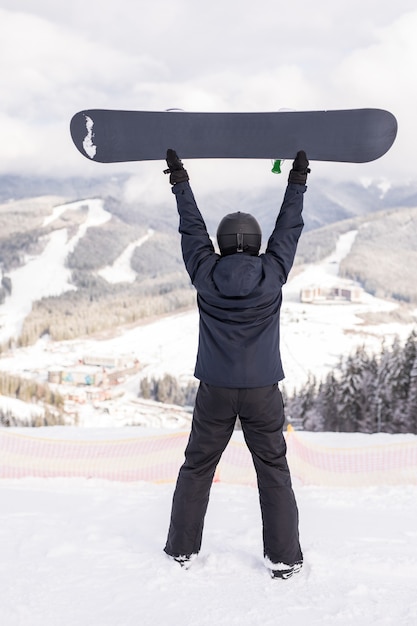 Photo happy man standing with snowboard over his head on the top of mountains hill