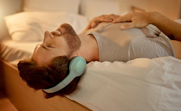 Happy man streaming a podcast in bed smiling with headphones at home young guy listening to music from spotify on internet or radio in earphones time to relax enjoy peace in his apartment bedroom