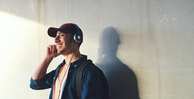 Happy man student and headphones listening to music on campus with happiness and audio against wall background Gen z education and male person at college mockup space and podcast streaming