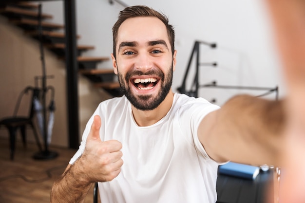 Happy man taking a selfie while sitting at the kitchen at home, peace gesture